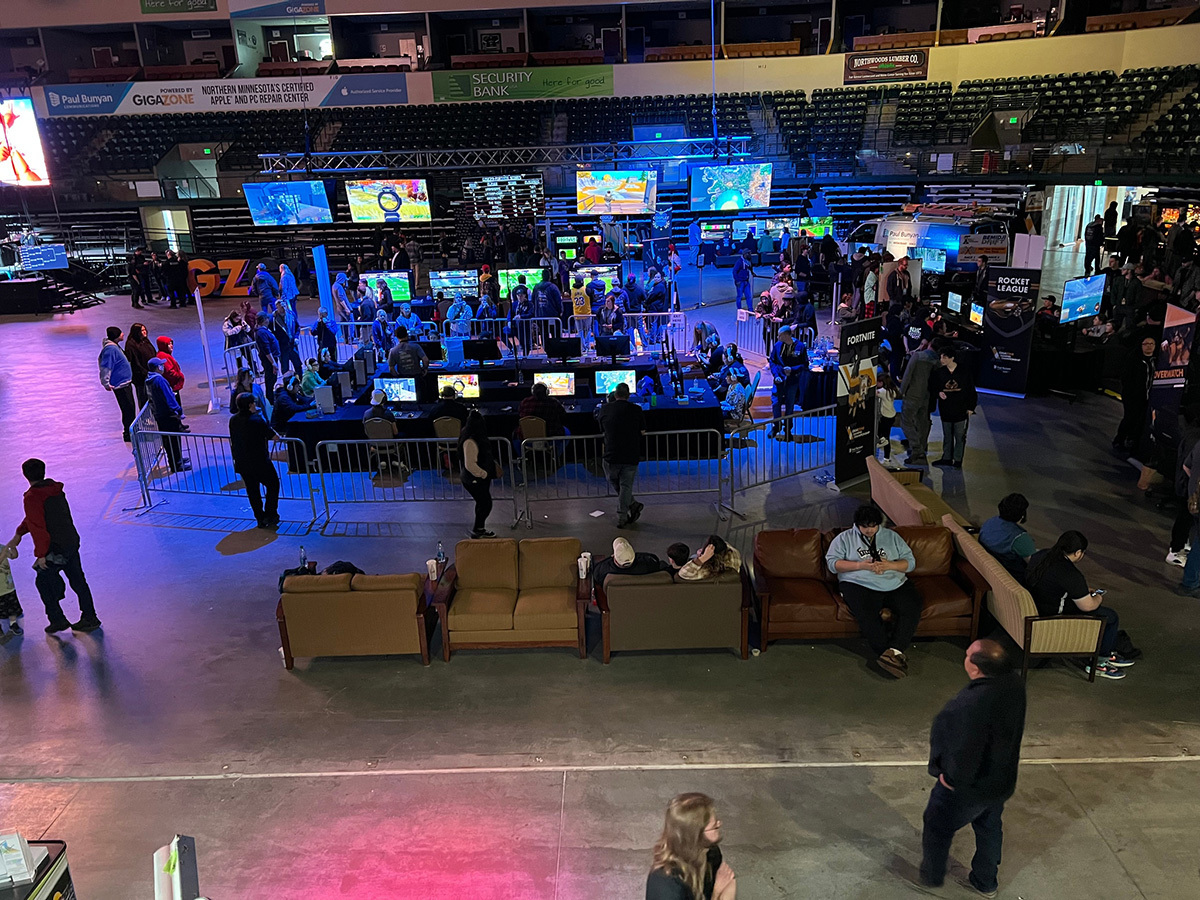 People walk around on an arena floor as others play video games at desks.