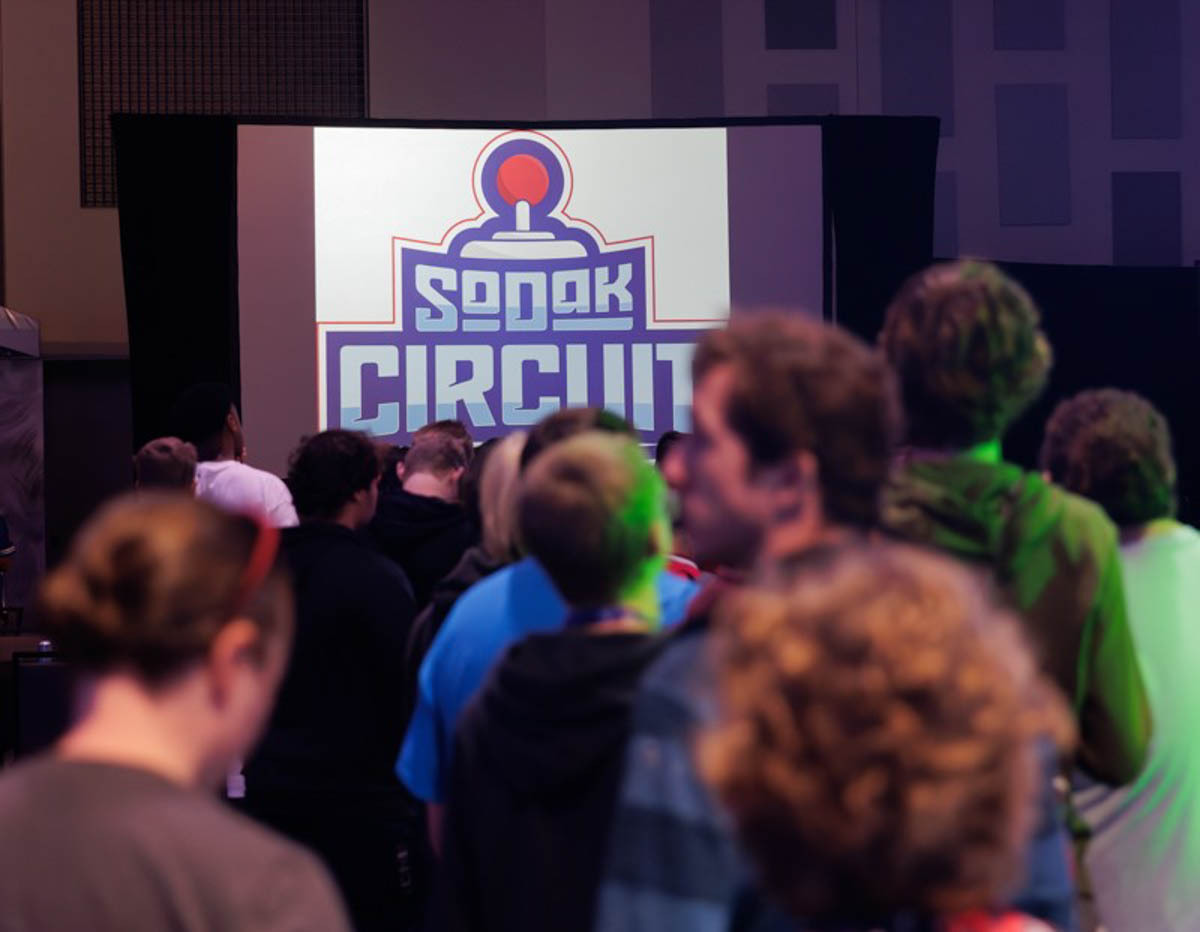 A crowd of people stand in line to register for the SoDak Circuit esports event with the SoDak Circuit logo on the screen in the background