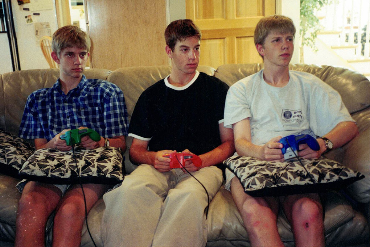 Three teenagers sit on a couch holding N64 controllers in the late 1990s