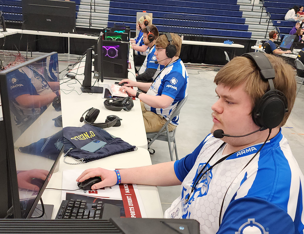 Esports players sit at a table in front of a bank of computers at a tournament.