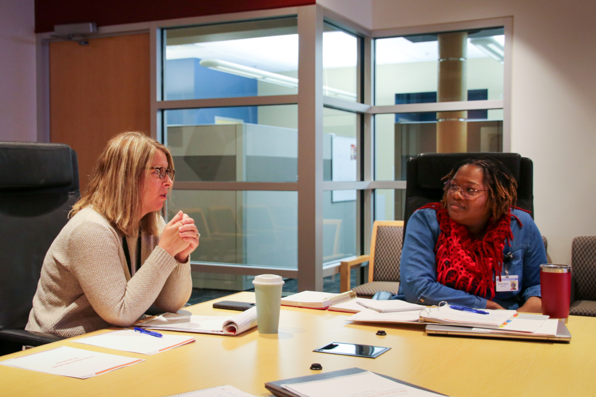 Jamie talks at the board table as Shamieko listens in