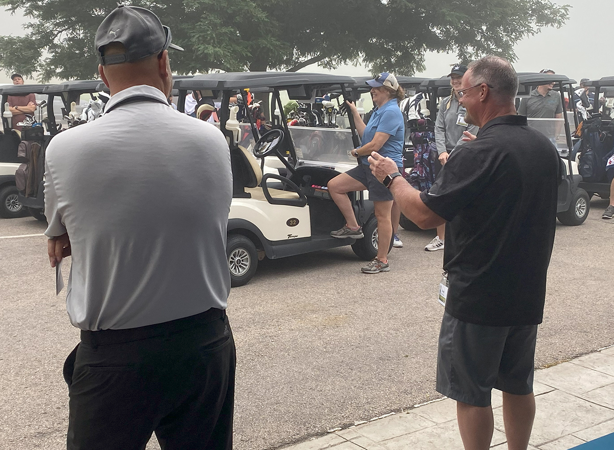 Two men in the foreground with the one on the right facing away from the camera and giving instructions to several golfers.