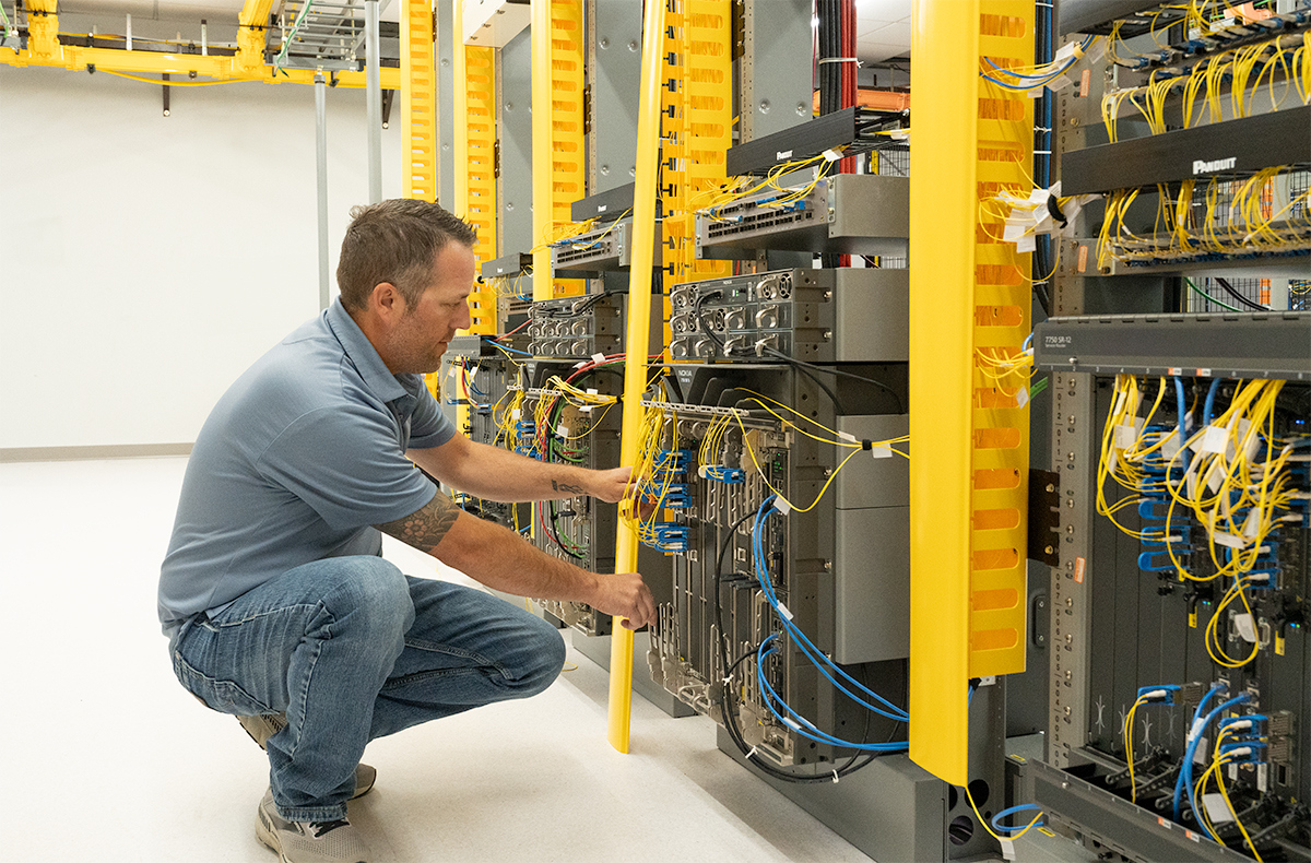 SDN Communications Network Engineer Riley Haag stands next to a wall of servers and cords in SDN's training lab.