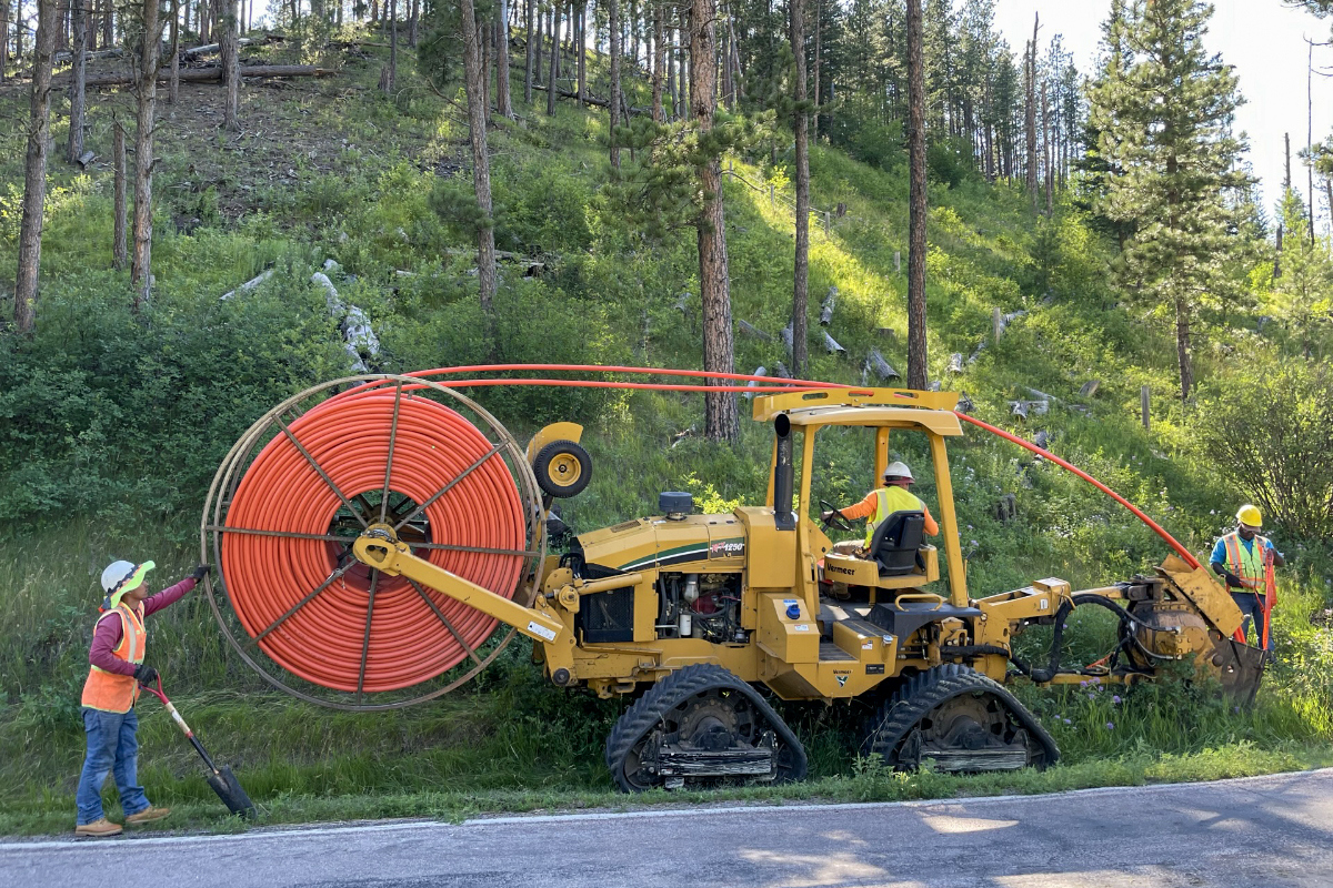 Crews using plow to cut fiber conduit into ground along a highway and against a steep hill