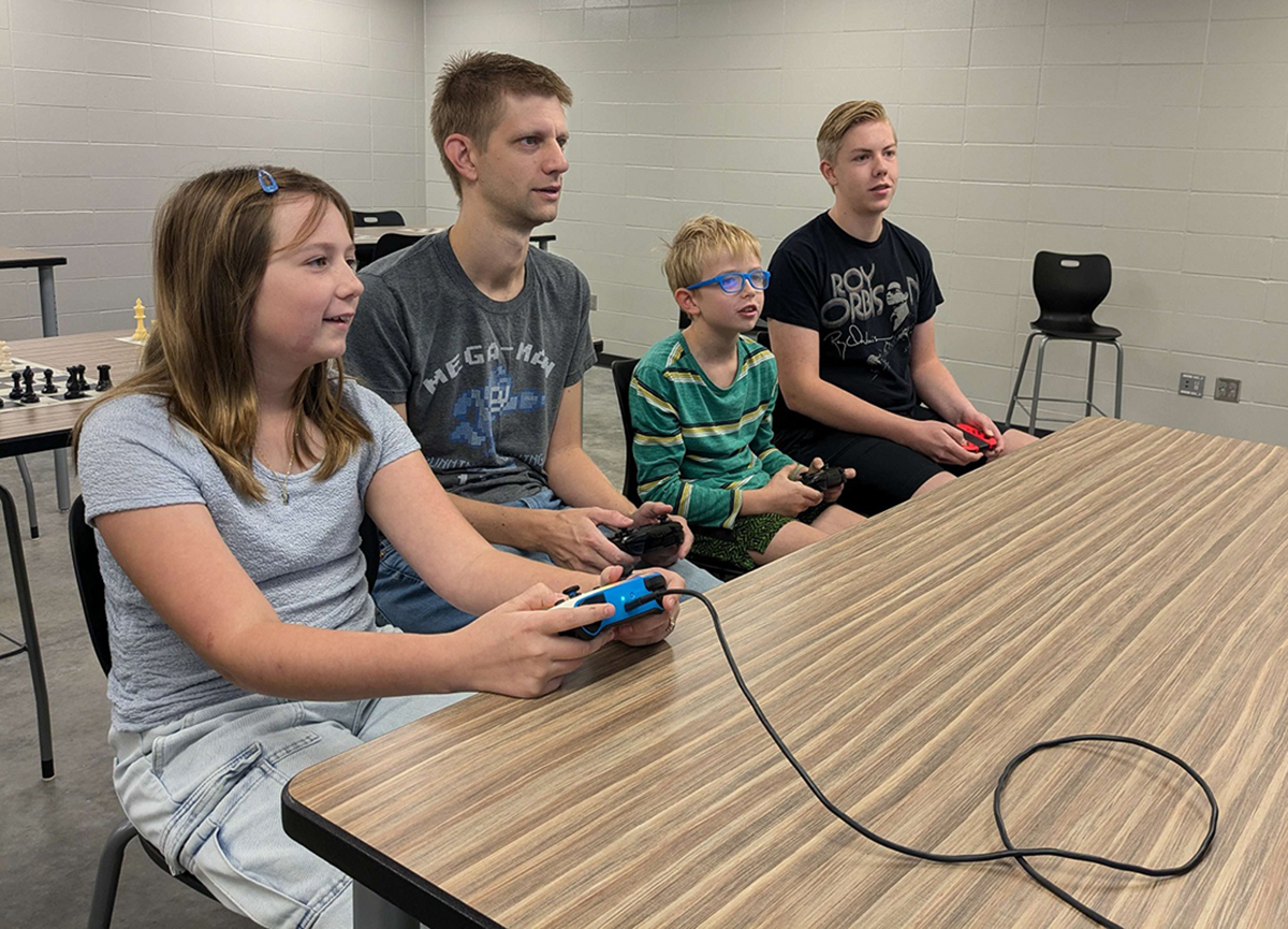 An adult and three kids sit at a table and watch a TV screen while playing Super Smash Bros. Ultimate.