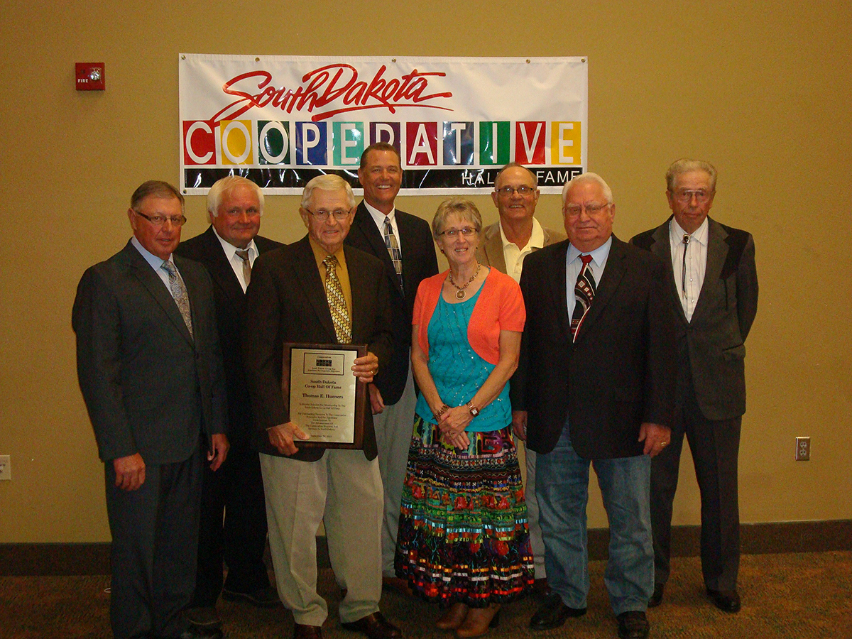 Eight people stand in front of a South Dakota Cooperative Hall of Fame sign and one person is holding a plaque.